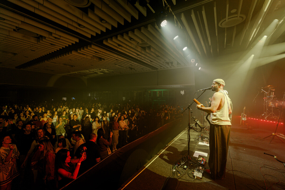 Show ao vivo em estúdio: músico cantando e tocando no palco com microfone, luzes de refletores e plateia lotada ao fundo, em apresentação musical