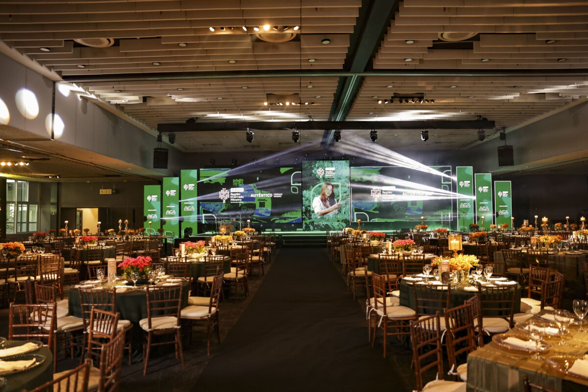 Salão de evento corporativo com corredor central, cadeiras de madeira e mesas decoradas com arranjos de flores, enquanto um telão com identidade verde apresenta programação e palestrante ao fundo.