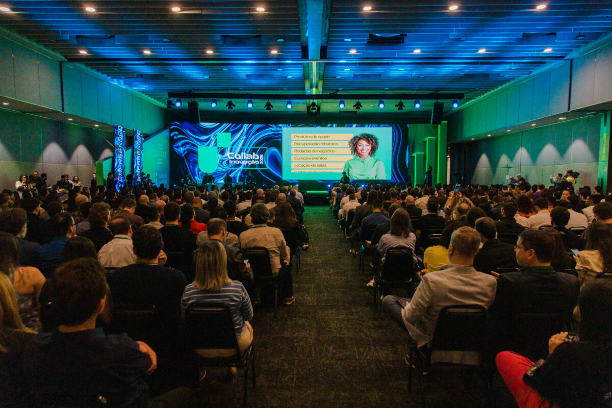 Auditório lotado durante evento corporativo, com palestrante ao centro do palco e telões digitais exibindo apresentação com gráficos e lista de tópicos. Iluminação em azul e verde.