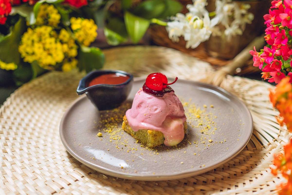 Sobremesa do Festival Sabores da Primavera, com uma preparação rosada coberta por calda de chocolate e finalizada com uma cereja, acompanhada por mais calda disposta ao lado.
