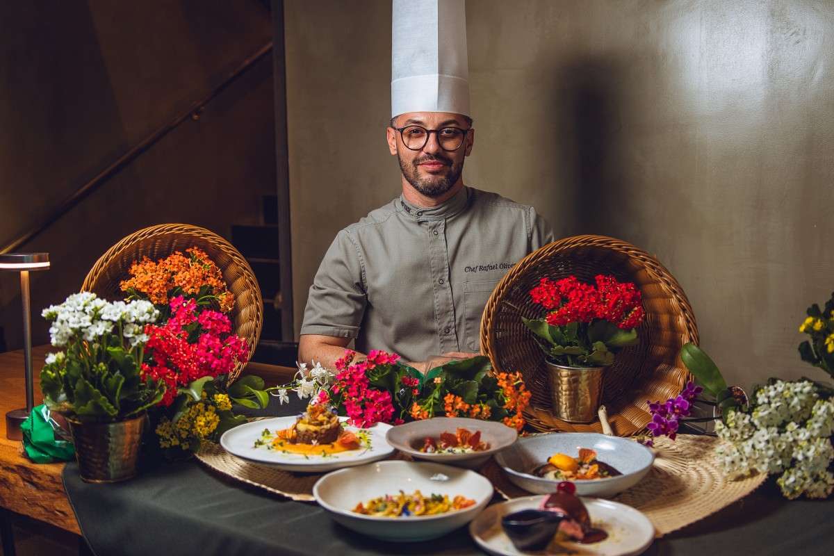 Chef Rafael Oliver apresenta os cinco pratos do Festival Sabores da Primavera, dispostos sobre uma mesa decorada com flores, folhagens e elementos naturais. As preparações têm diferentes cores, texturas e finalizações.