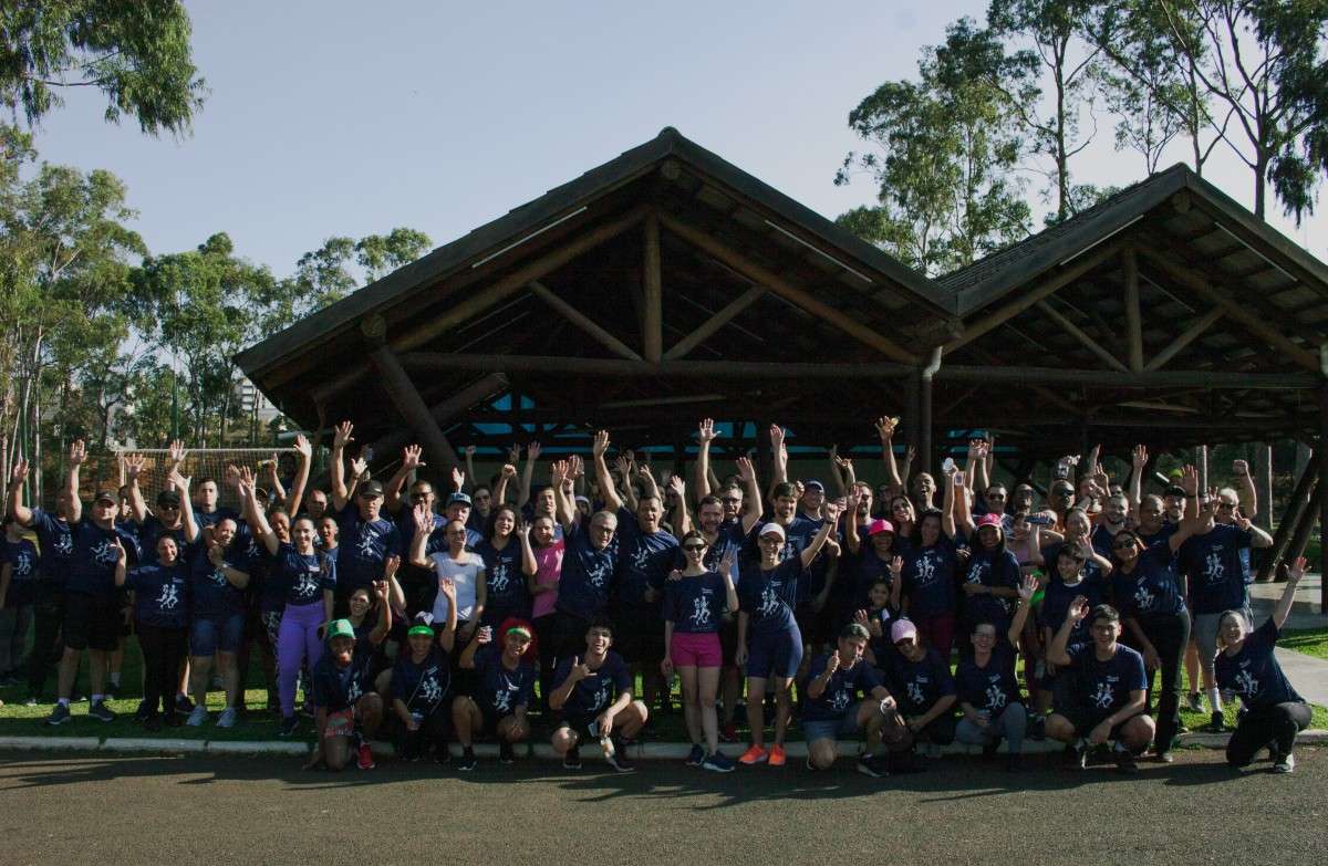 Participantes da caminhada solidária da EPS posam juntos com os braços erguidos no Parque do Sabiá, diante de um quiosque e cercados por árvores.