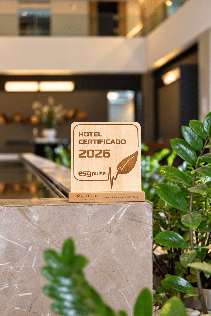 placa oficial enviada pela ESG Pulse escrita “Hotel Certificado 2026 ESG Pulse” e o nome Mercure Uberlândia Plaza Shopping, exposta sobre um balcão de mármore no térreo da Torre 2, em ambiente interno com plantas.