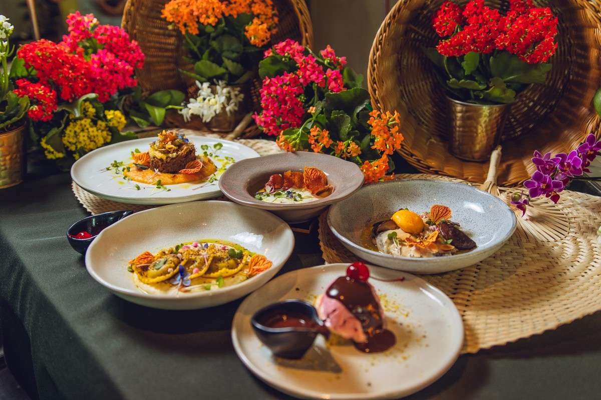 Cinco pratos do Festival Sabores da Primavera dispostos sobre uma mesa: quatro preparações salgadas e uma sobremesa em primeiro plano, cercadas por flores vermelhas, laranjas, amarelas e roxas.
